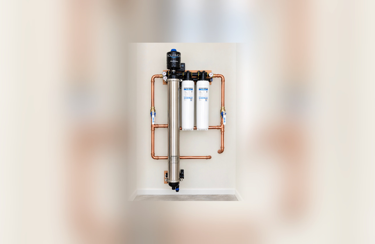 A Heritage suite installed on a utility room wall: a tall stainless ultrafiltration vessel with a black Aquamor cap, two white carbon cartridge housings beside it, pressure gauges above, and polished copper pipework running in and out through isolation valves.